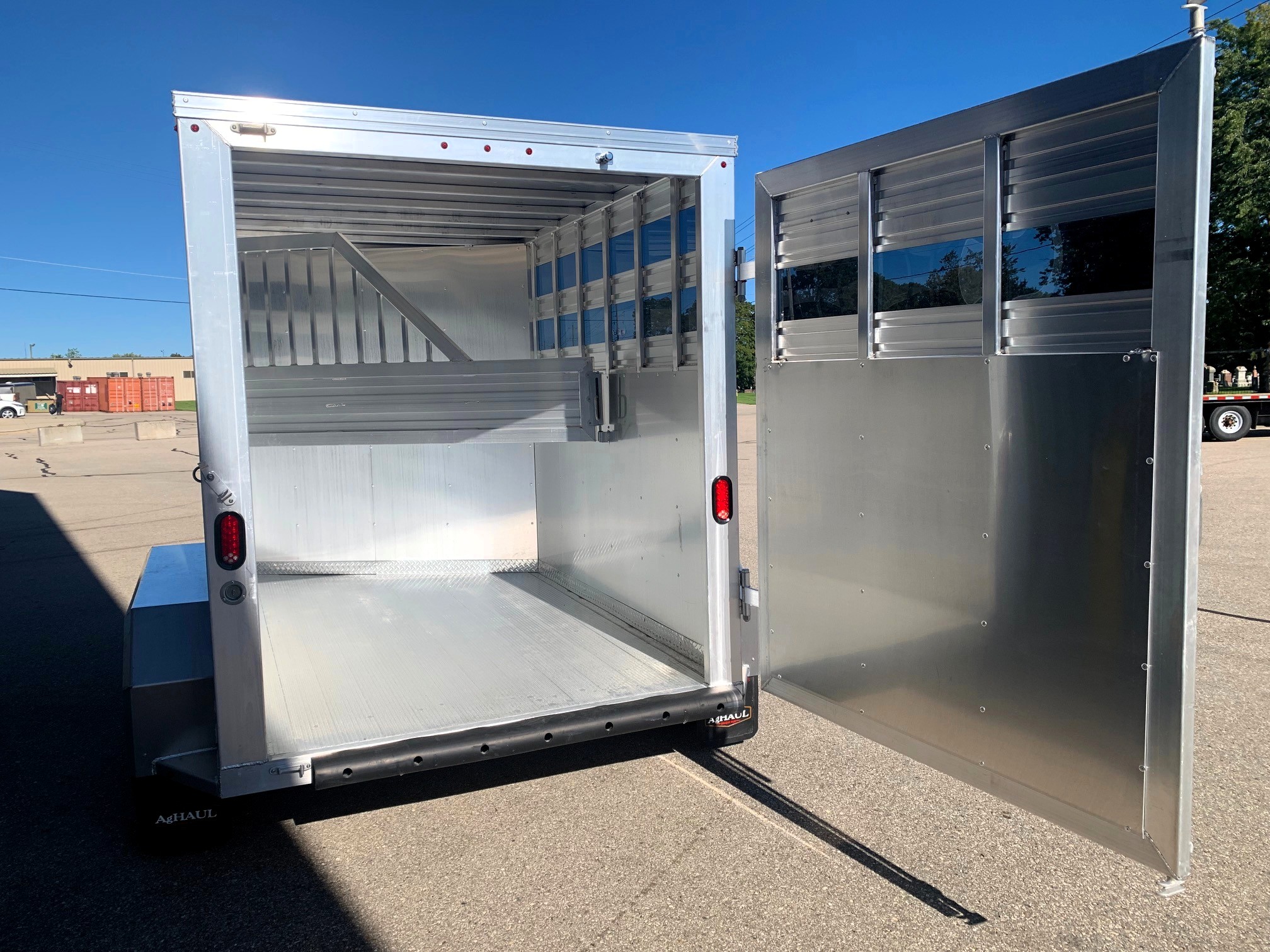 image6 40 - McFarlane Trailer Sales Open aluminum trailer with rear ramp lowered, showing interior divider and ribbed walls against a bright blue sky.