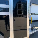 image8 16 - McFarlane Trailer Sales Rear corner of a shiny aluminum trailer with a large black glass door framed by metal, featuring a rounded U-shaped handle and diamond-plate trim along the edge; small orange marker light near the corner.