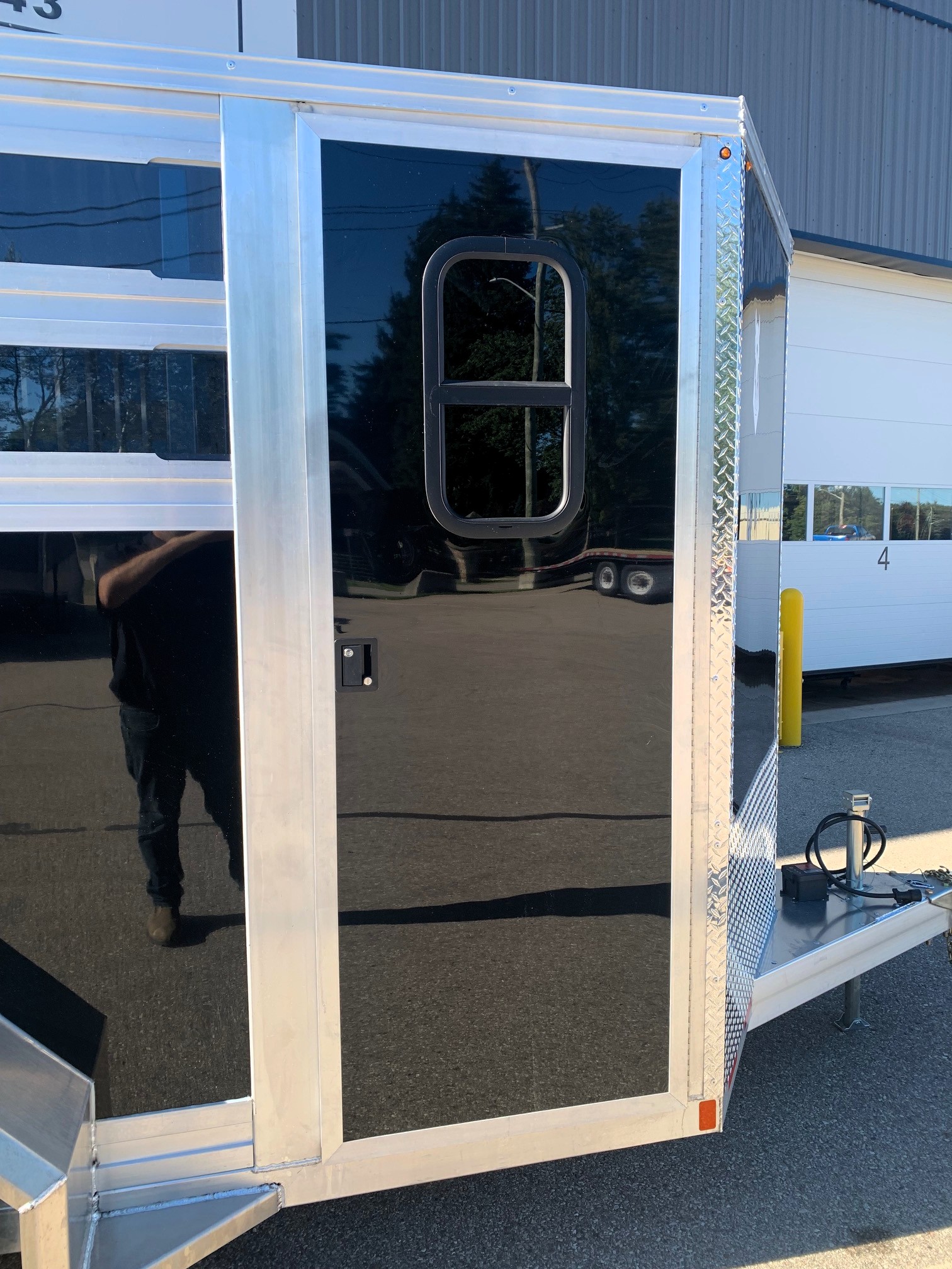 image8 16 - McFarlane Trailer Sales Rear corner of a shiny aluminum trailer with a large black glass door framed by metal, featuring a rounded U-shaped handle and diamond-plate trim along the edge; small orange marker light near the corner.