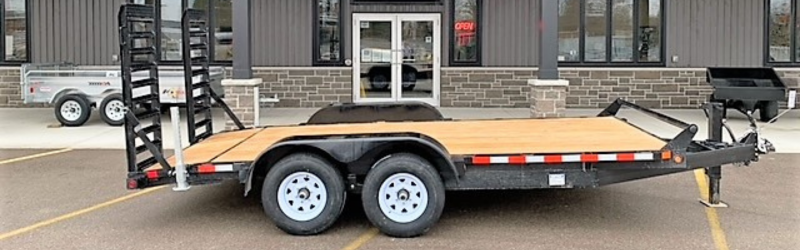 Dual-axle flatbed trailer with a wooden deck and rear loading ramps parked in a parking lot in front of a commercial building.