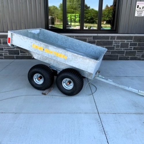 Galvanized utility trailer with two axles and a tipped bed, attached to a tow bar, parked on a concrete pad in front of a building.
