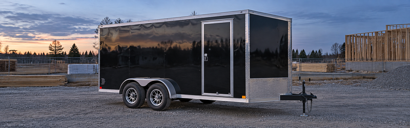 Black enclosed tandem-axle cargo trailer on a gravel lot at dusk, with a construction site in the background.